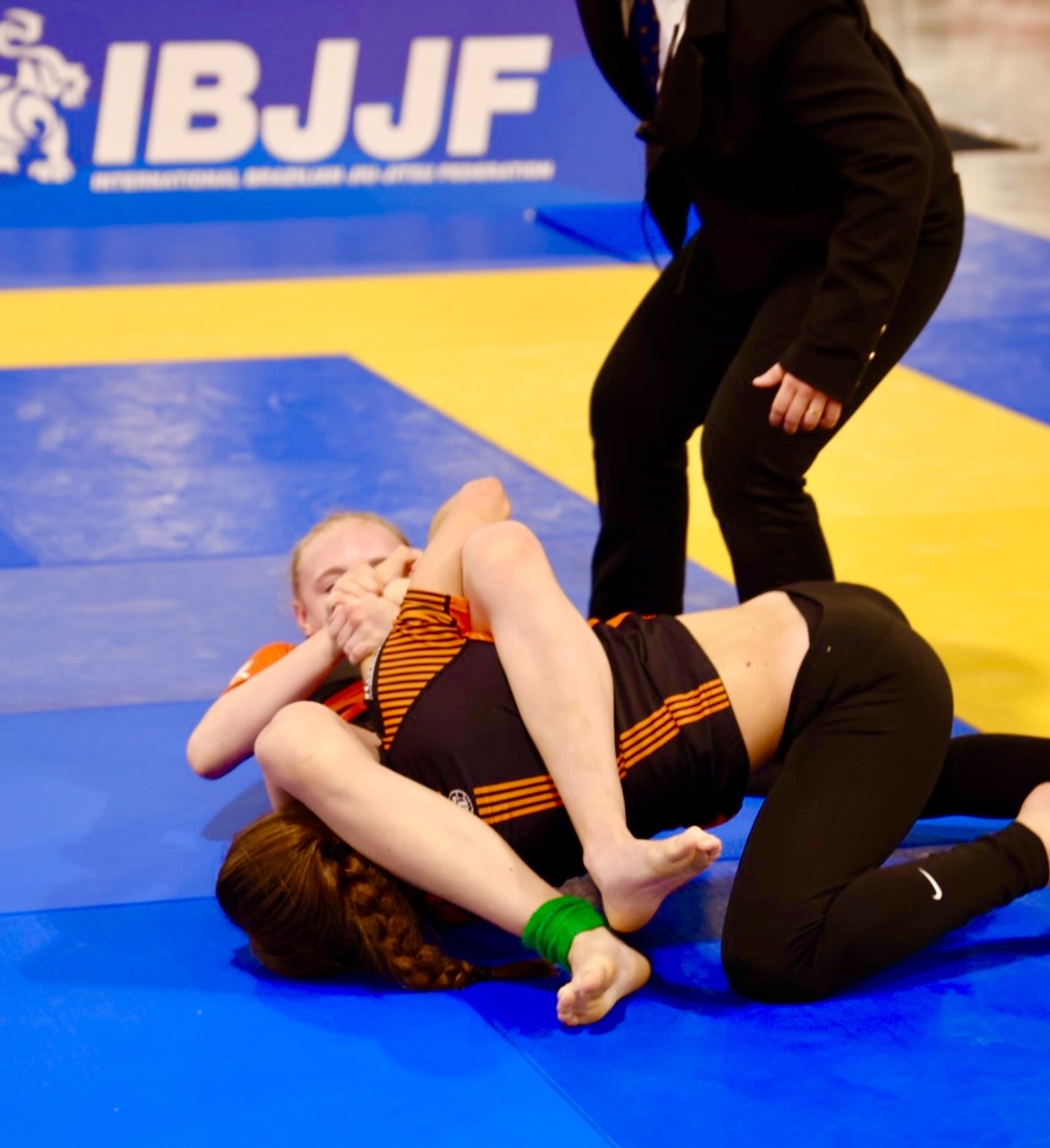 Weiteres Wettkampfbild aus dem Turnierbaum der IBJJF Pan Kids No-Gi Championship 2024. Die abgebildete Person befindet sich in einem Übergang von der Kontrolle zur Submission. Auf der Rückwand der Wettkampfmatte ist IBJJF-Branding sichtbar.